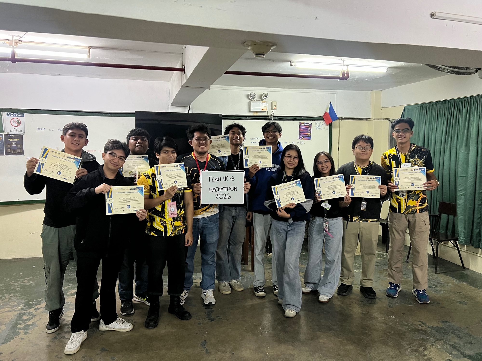 Team UC-B holding their University of Cebu certificates after the 2026 hackathon, with a whiteboard reading TEAM UC-B HACKATHON 2026